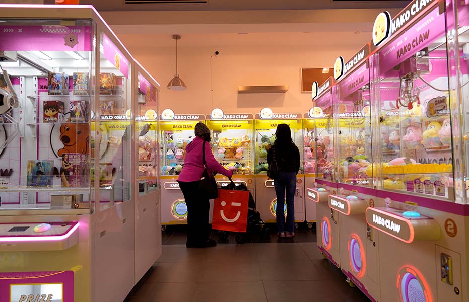 Two guests browsing the selection of prize machines at a brightly lit Kako Claw store.